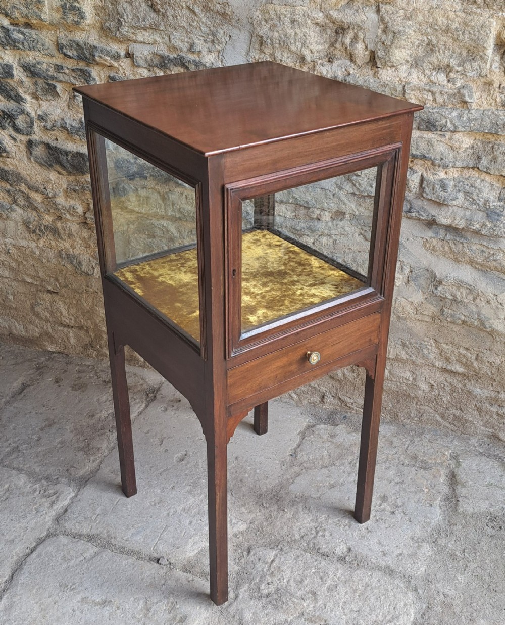A Georgian mahogany standing display cabinet featuring a glass-enclosed upper section lined with gold fabric, a single lower drawer, and square tapered legs. View II