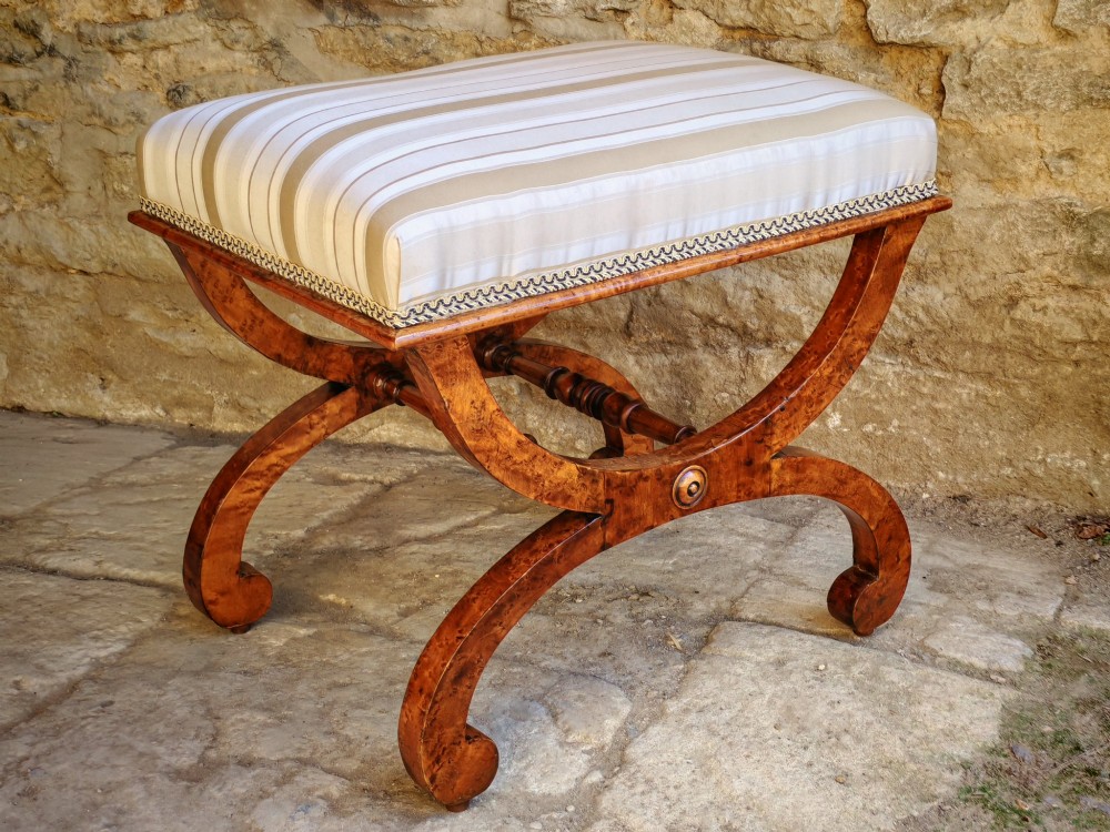 A mid-19th century X-frame stool crafted from figured burr maple, featuring a central turned stretcher and a neutral striped fabric upholstered seat.