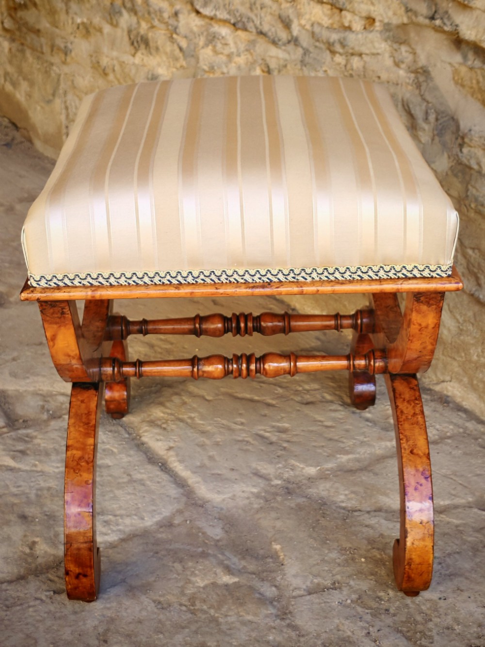 A mid-19th century X-frame stool crafted from figured burr maple, featuring a central turned stretcher and a neutral striped fabric upholstered seat. View V