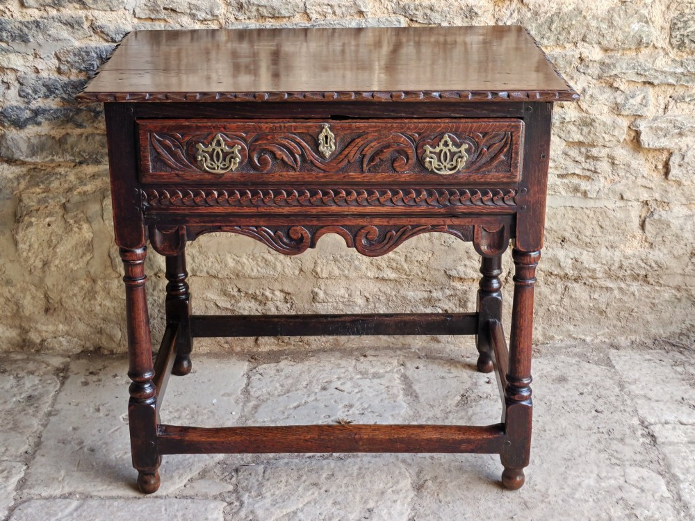 An 18th-century dark oak lowboy table featuring a hand-carved drawer front, decorative brass handles, turned legs, and a lower stretcher base.