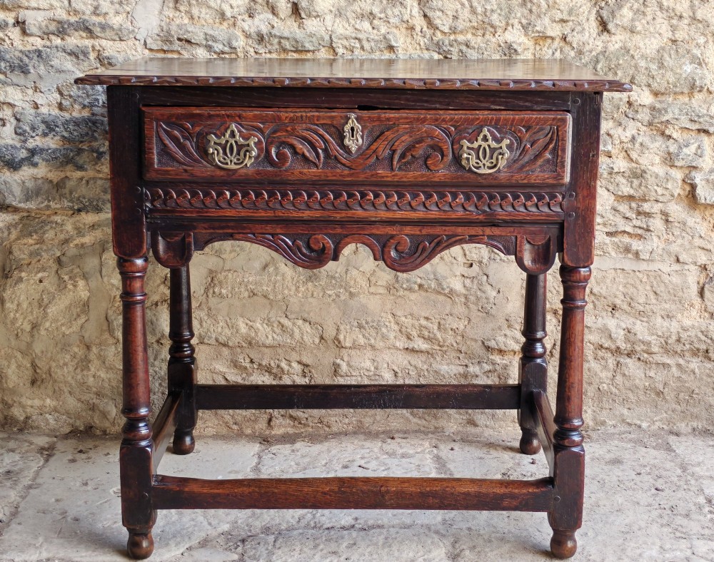 An 18th-century dark oak lowboy table featuring a hand-carved drawer front, decorative brass handles, turned legs, and a lower stretcher base. View II