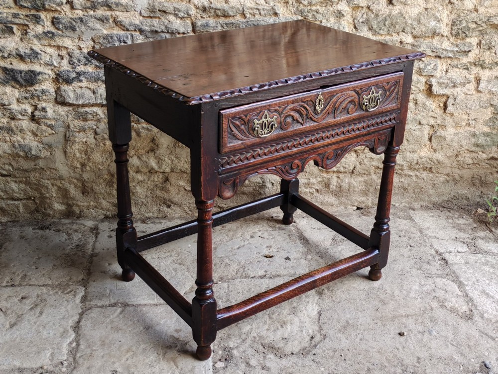 An 18th-century dark oak lowboy table featuring a hand-carved drawer front, decorative brass handles, turned legs, and a lower stretcher base. View VI