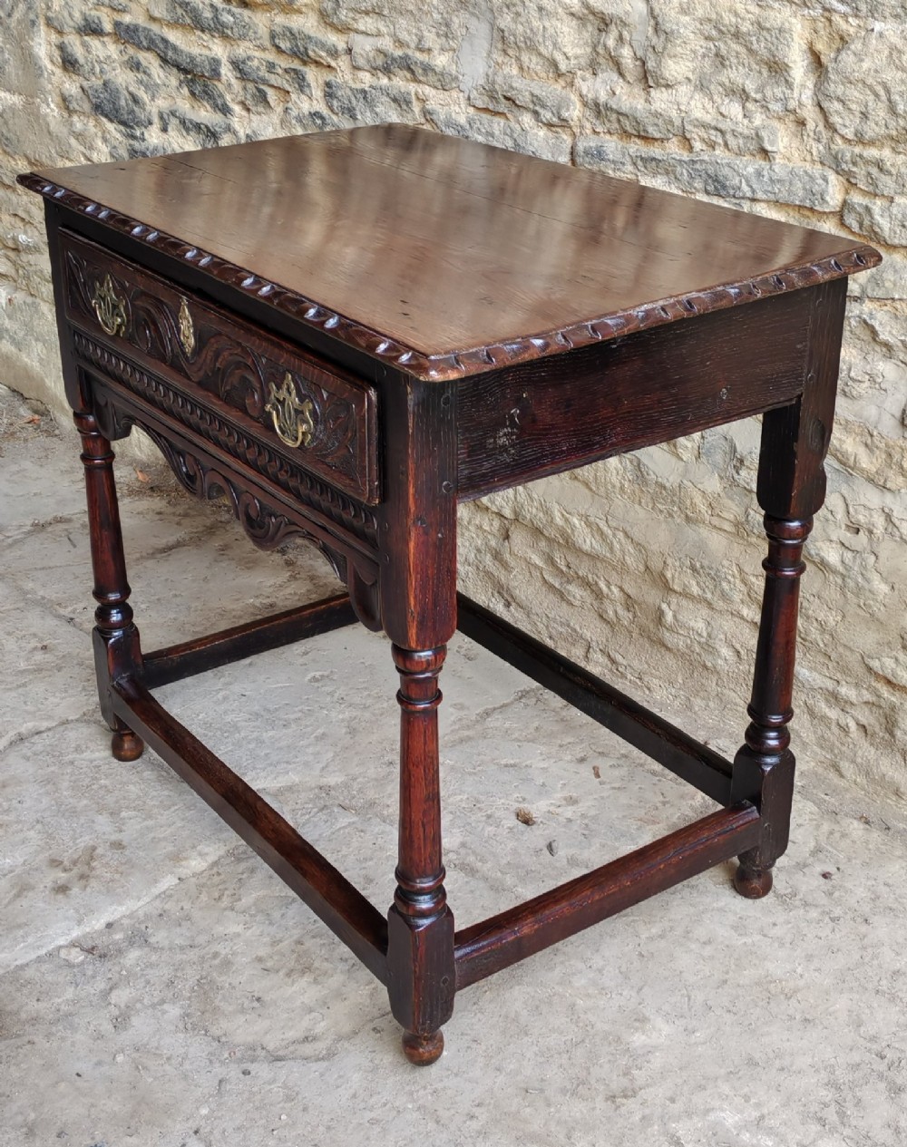 An 18th-century dark oak lowboy table featuring a hand-carved drawer front, decorative brass handles, turned legs, and a lower stretcher base. View VII