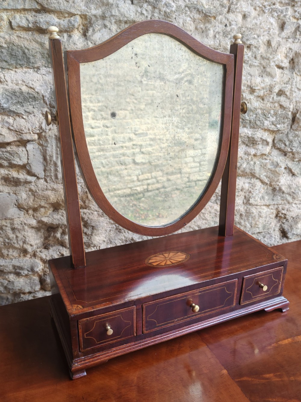 A Georgian mahogany dressing mirror featuring a shield-shaped swivel frame, bone finials, and an inlaid three-drawer box base.