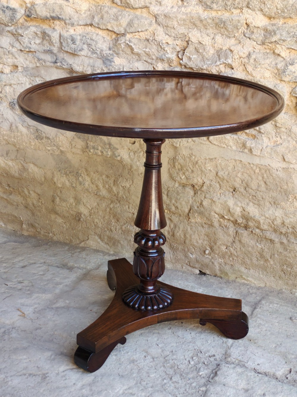 A Regency period dark mahogany and rosewood circular dish top occasional table resting on a carved pedestal stem and a triform base. View VIII
