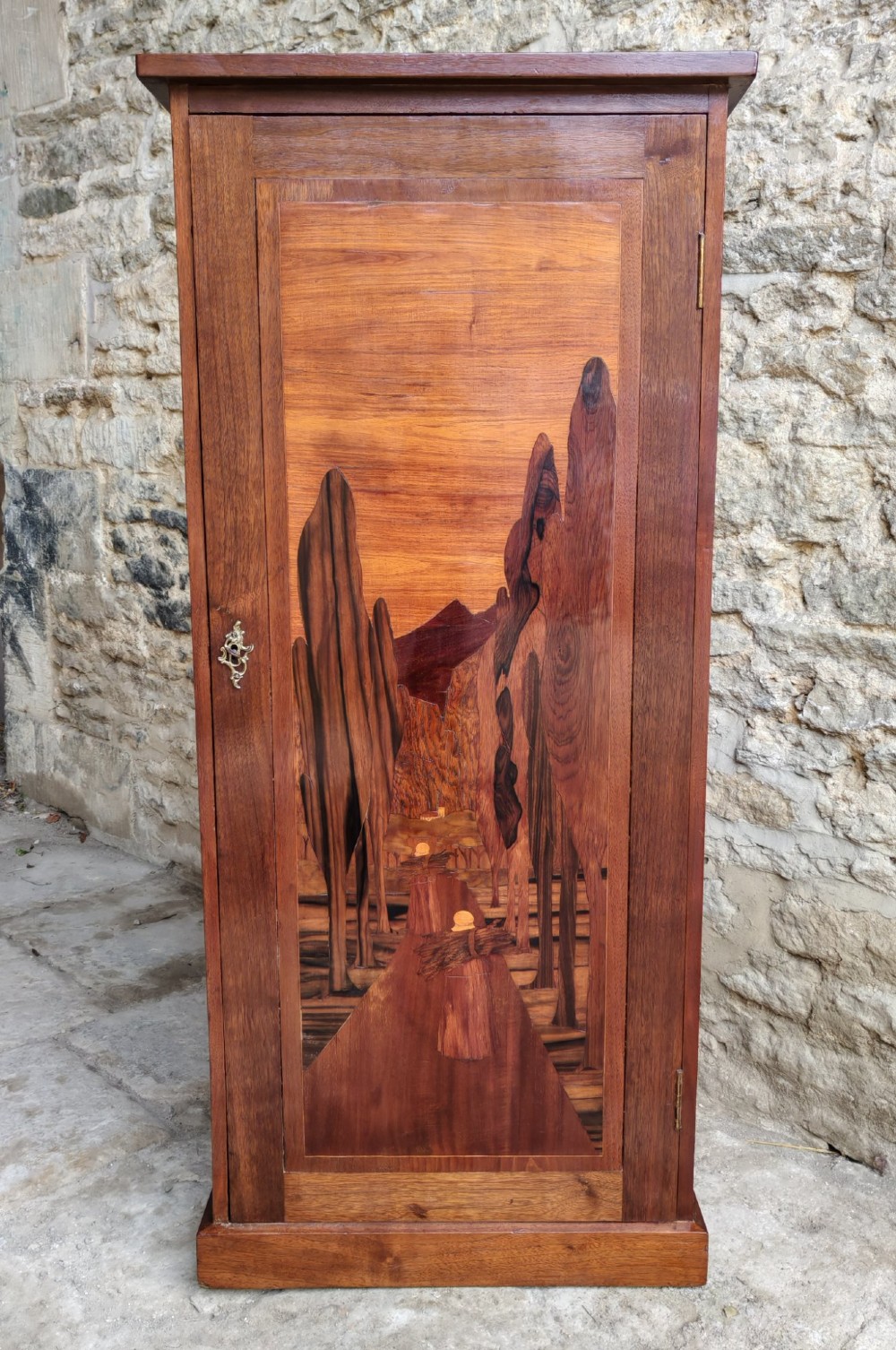 A tall mahogany single-door cupboard featuring a prominent marquetry inlaid panel depicting a mountain landscape with tall trees and a central path.