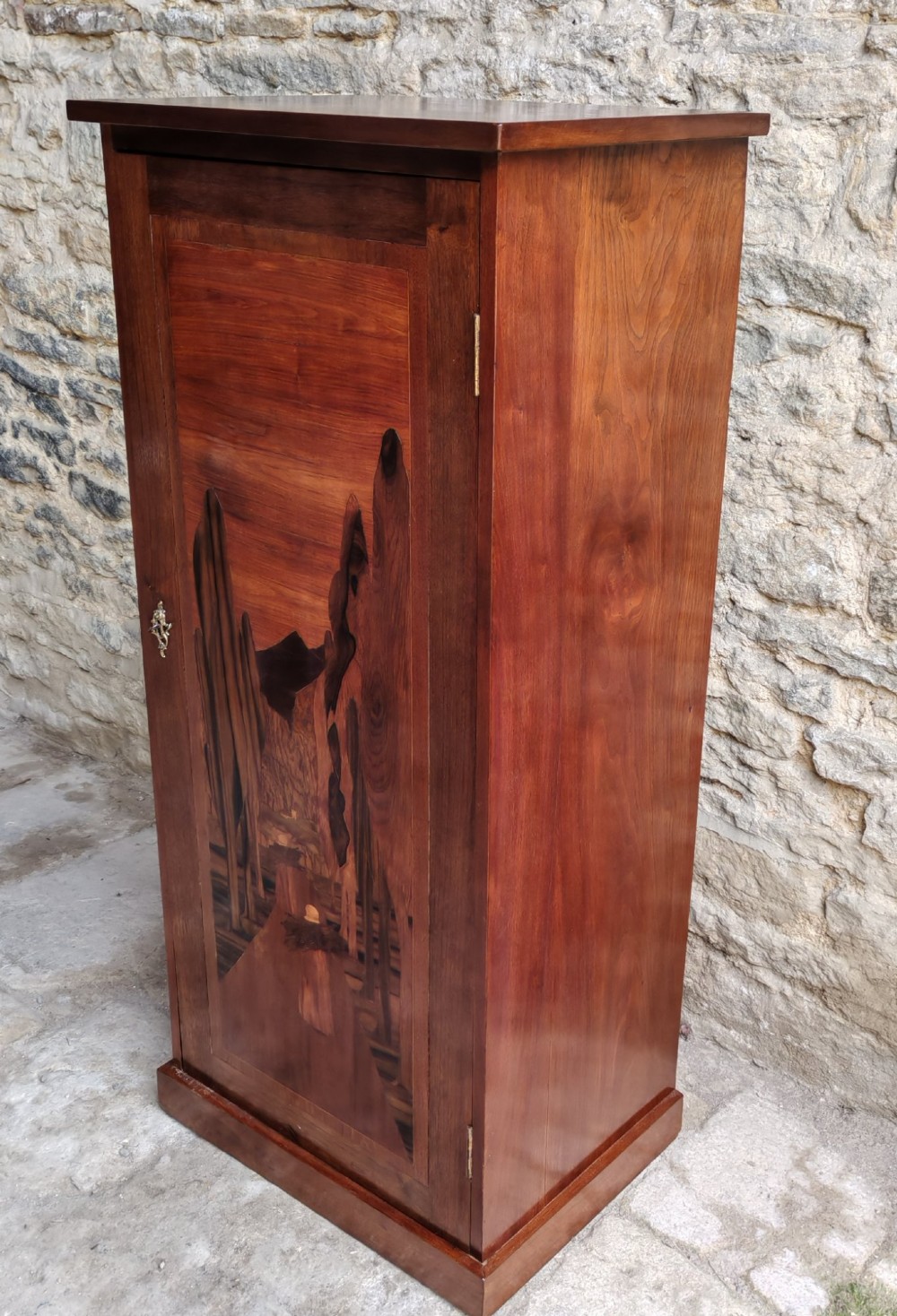 A tall mahogany single-door cupboard featuring a prominent marquetry inlaid panel depicting a mountain landscape with tall trees and a central path. View II