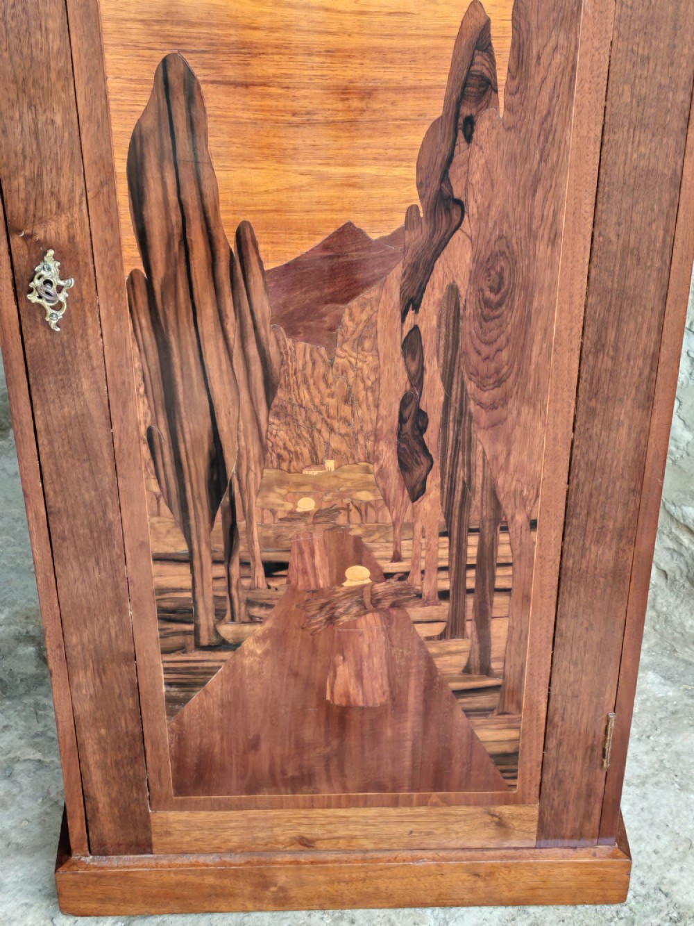A tall mahogany single-door cupboard featuring a prominent marquetry inlaid panel depicting a mountain landscape with tall trees and a central path. View V