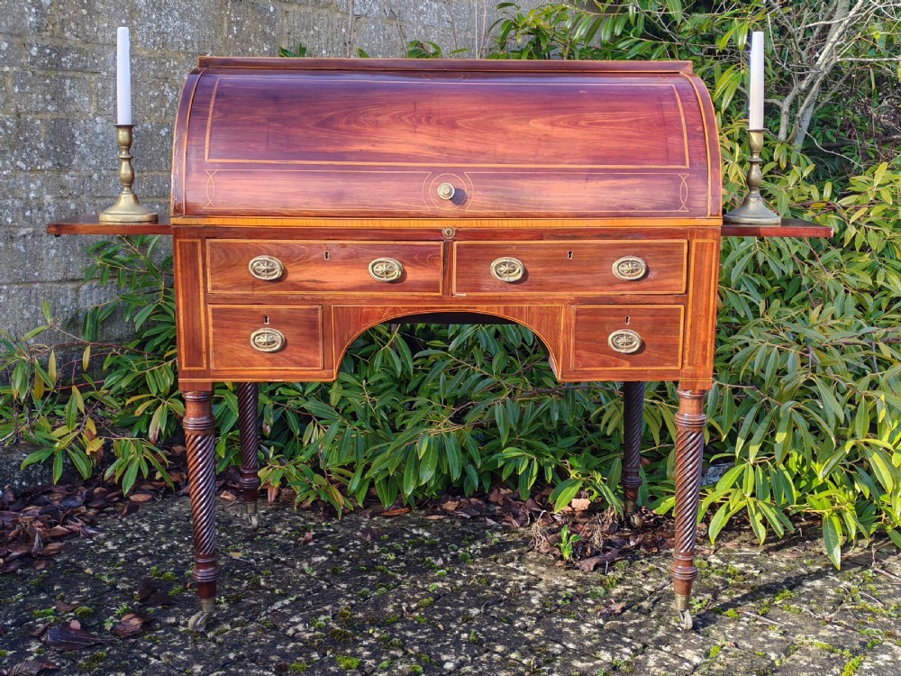 A Georgian mahogany cylinder top desk featuring a curved roll lid, inlaid detailing, multiple drawers with brass handles, pull-out side slides, and spiral turned legs on brass castors.