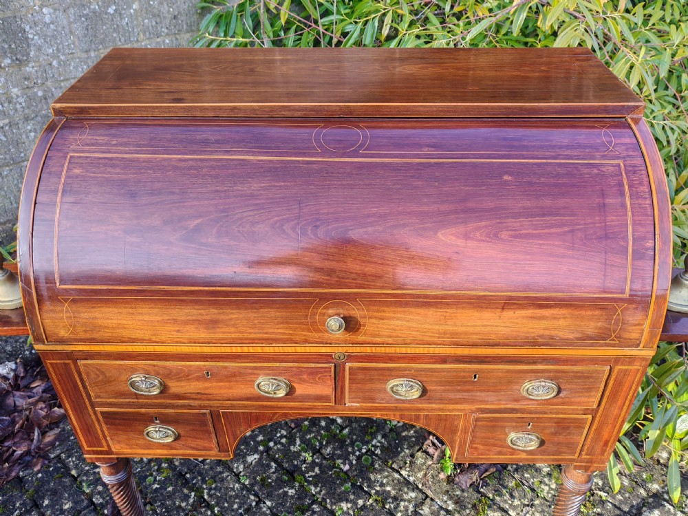 A Georgian mahogany cylinder top desk featuring a curved roll lid, inlaid detailing, multiple drawers with brass handles, pull-out side slides, and spiral turned legs on brass castors. View II