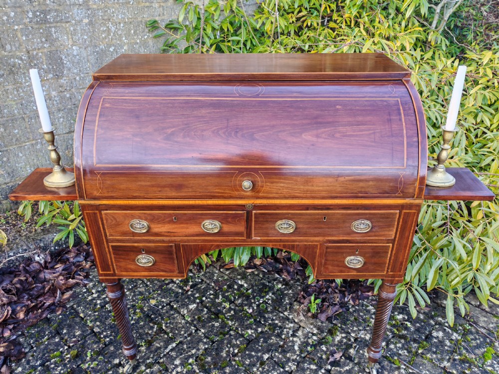 A Georgian mahogany cylinder top desk featuring a curved roll lid, inlaid detailing, multiple drawers with brass handles, pull-out side slides, and spiral turned legs on brass castors. View III