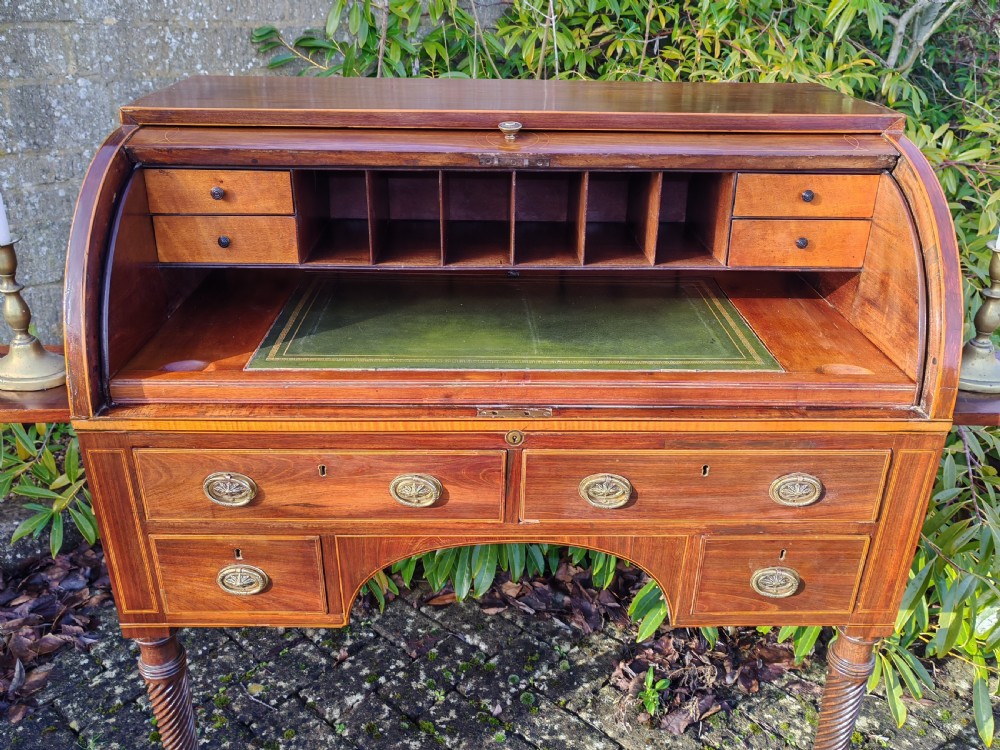 A Georgian mahogany cylinder top desk featuring a curved roll lid, inlaid detailing, multiple drawers with brass handles, pull-out side slides, and spiral turned legs on brass castors. View IV