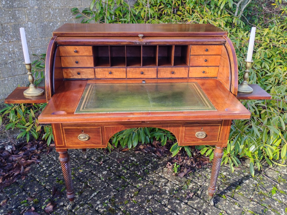 A Georgian mahogany cylinder top desk featuring a curved roll lid, inlaid detailing, multiple drawers with brass handles, pull-out side slides, and spiral turned legs on brass castors. View V