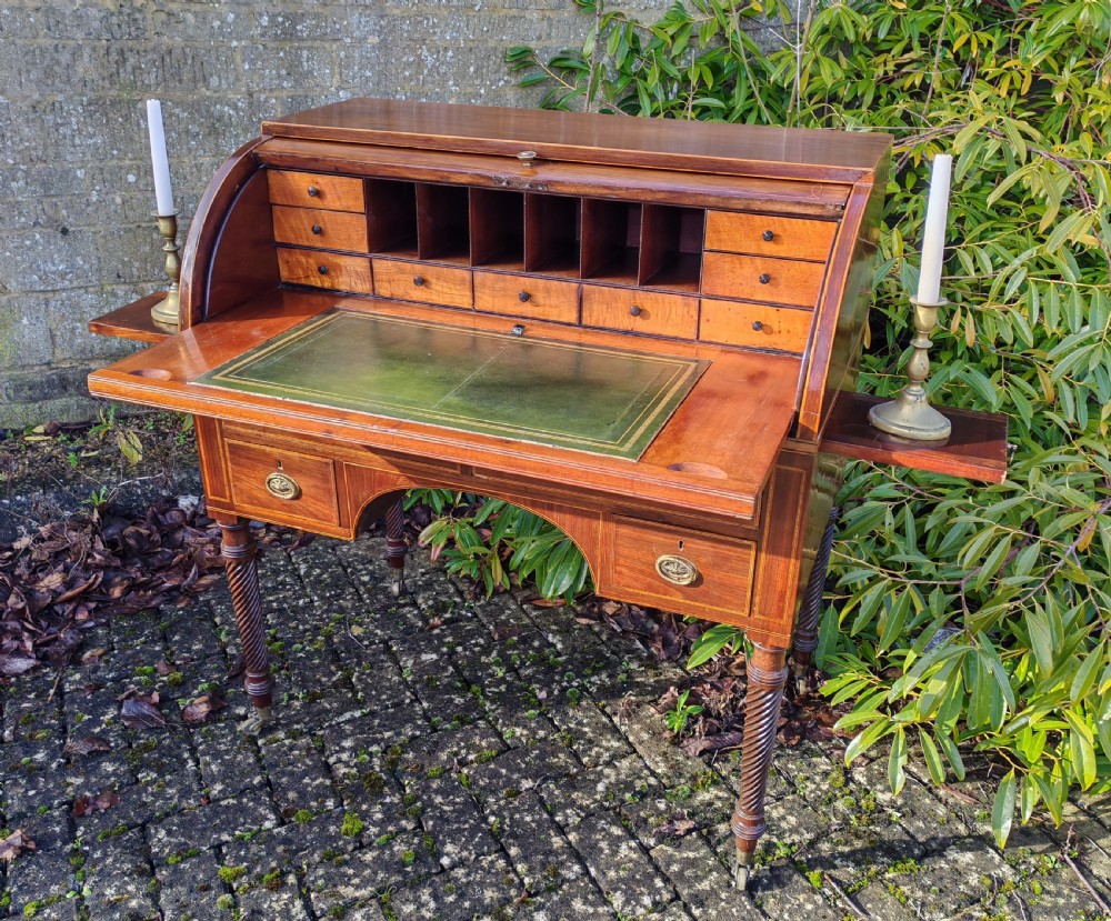 A Georgian mahogany cylinder top desk featuring a curved roll lid, inlaid detailing, multiple drawers with brass handles, pull-out side slides, and spiral turned legs on brass castors. View VI