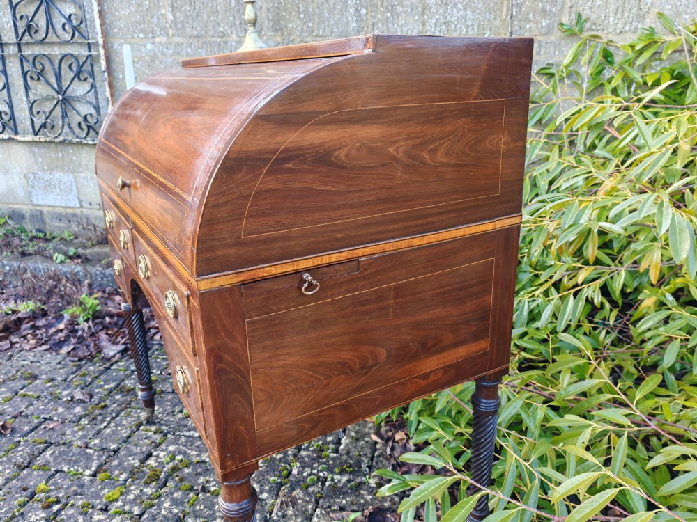 A Georgian mahogany cylinder top desk featuring a curved roll lid, inlaid detailing, multiple drawers with brass handles, pull-out side slides, and spiral turned legs on brass castors. View VIII