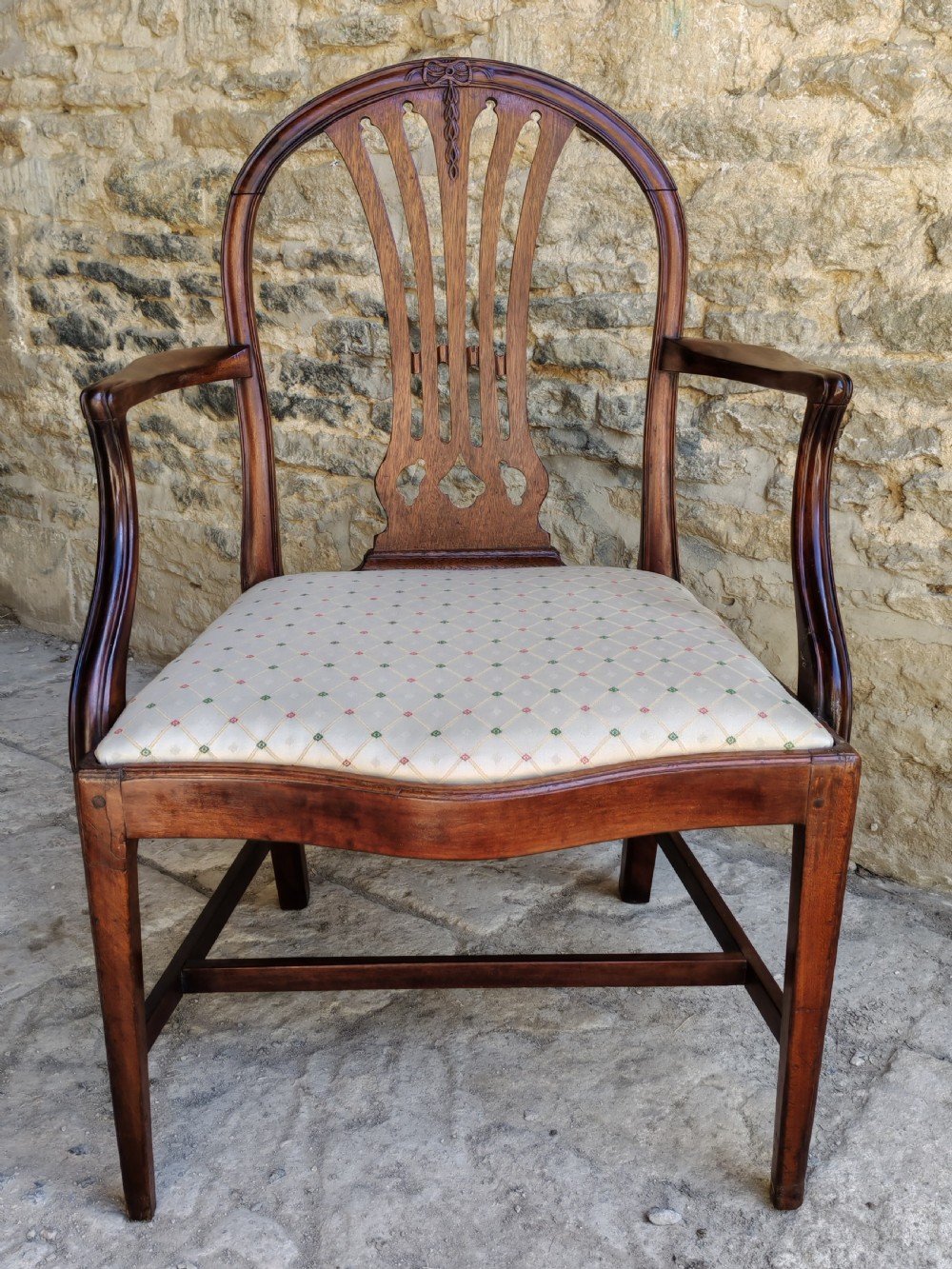 An 18th-century mahogany Hepplewhite carver armchair featuring an arched back with a pierced splat, sweeping armrests, an patterned upholstered seat, and square tapered legs.