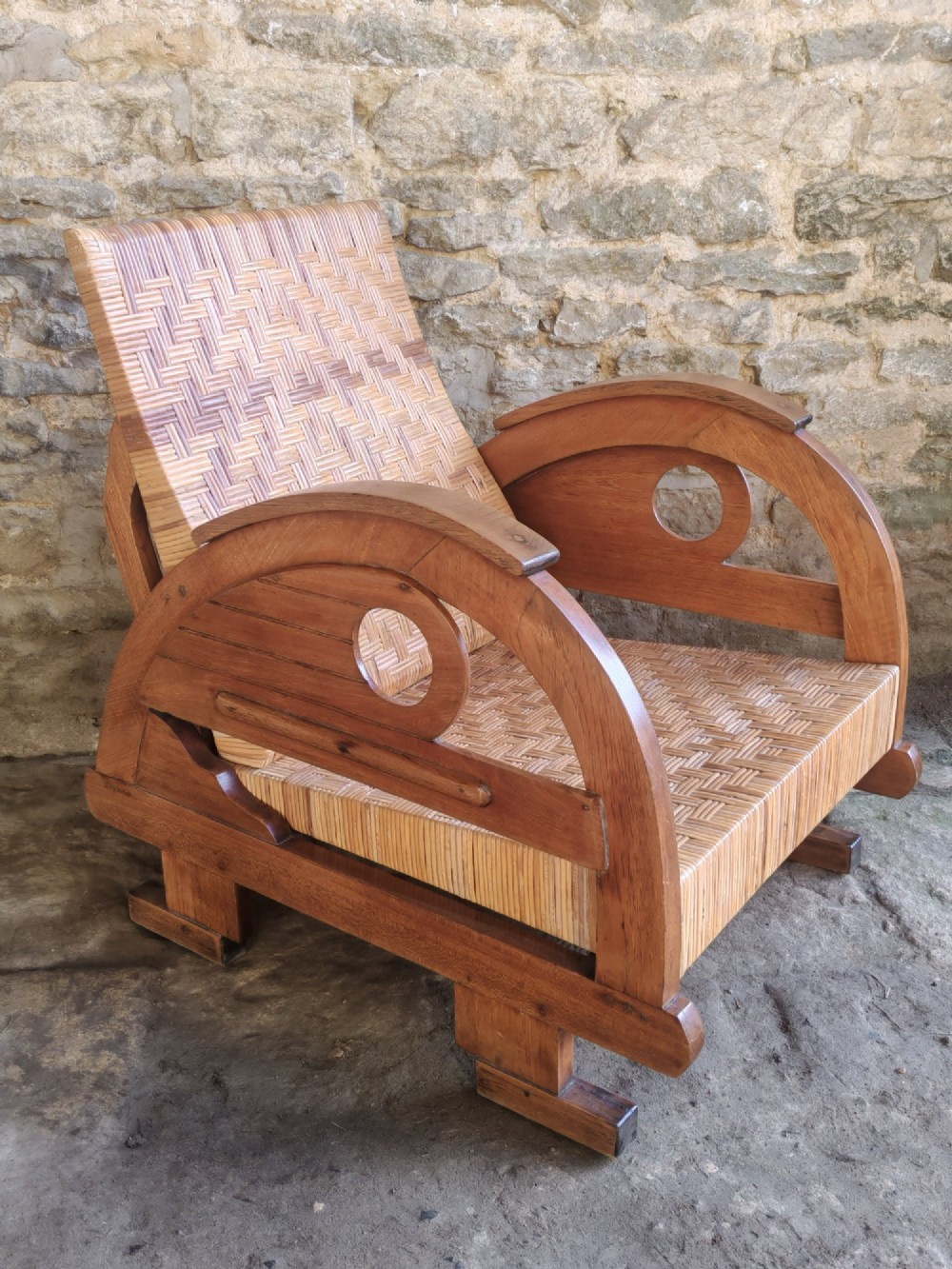 An Art Deco low-slung armchair featuring thick curved wooden armrests with circular cut-outs and a woven rattan seat and backrest.