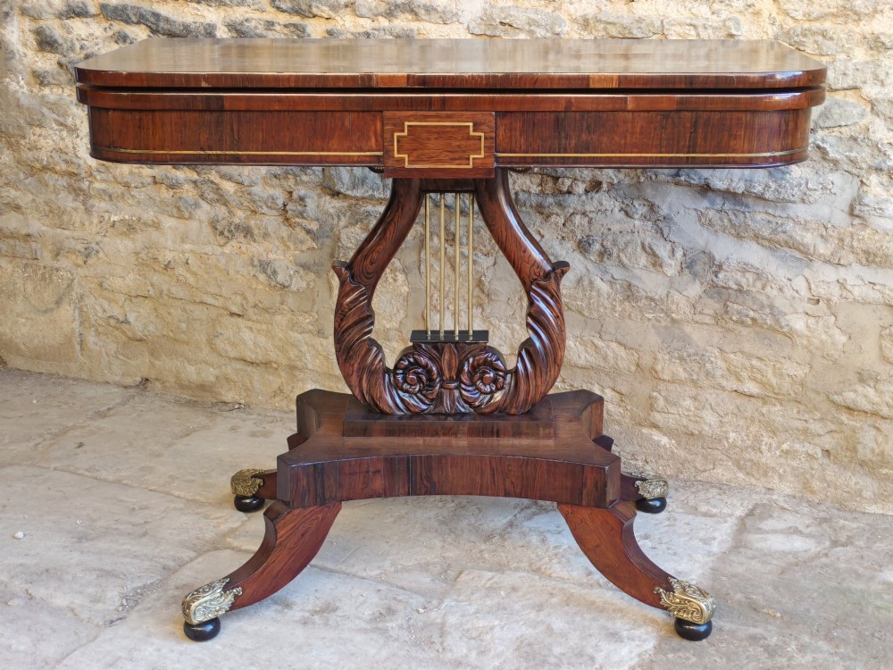 A Regency period rosewood tea table featuring a fold-over top, a central lyre-shaped support with brass rods, and a four-footed pedestal base with brass castors.