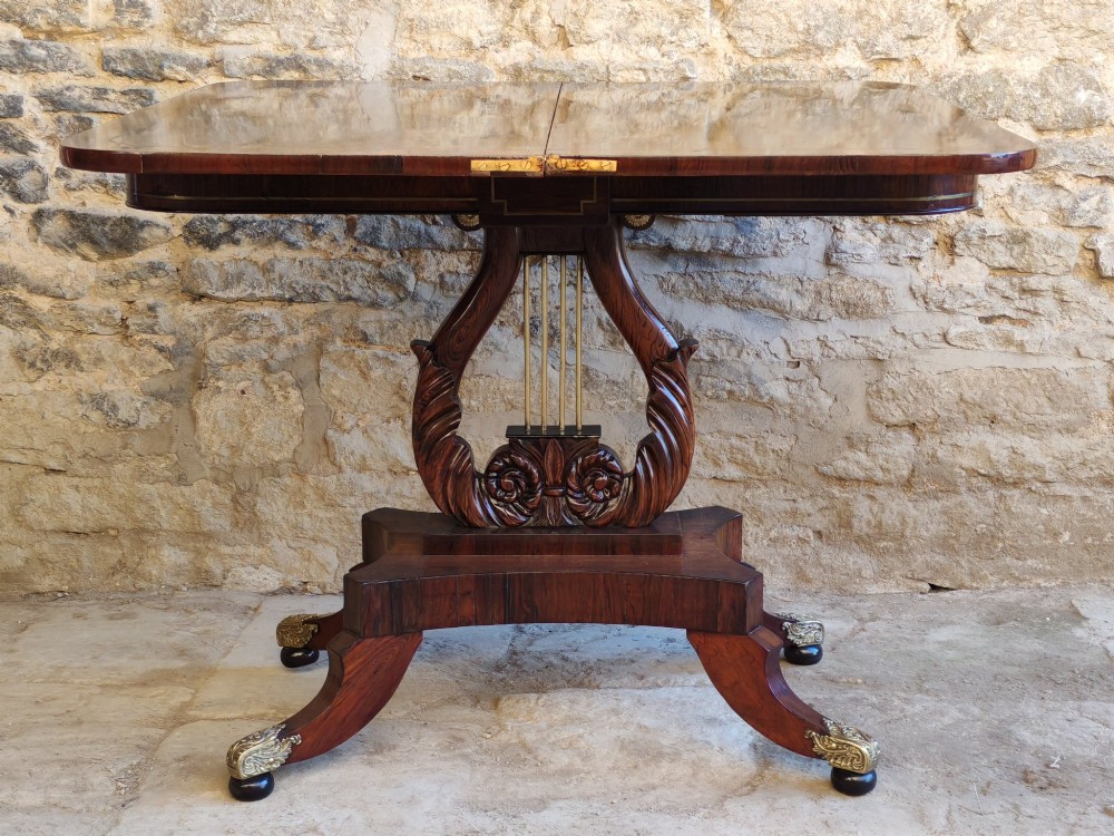 A Regency period rosewood tea table featuring a fold-over top, a central lyre-shaped support with brass rods, and a four-footed pedestal base with brass castors. View X