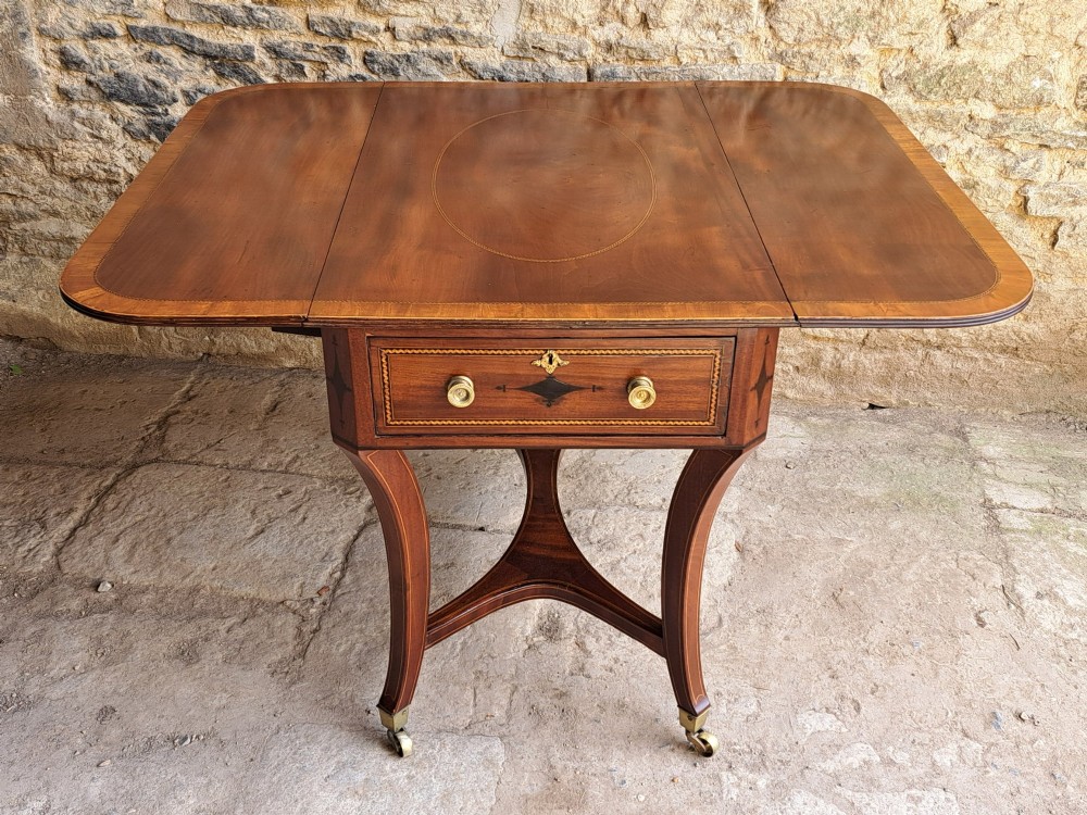 A Regency mahogany drop-leaf tea table featuring satinwood crossbanding, an oval marquetry motif, a single drawer with brass knobs, and splayed legs on castors.