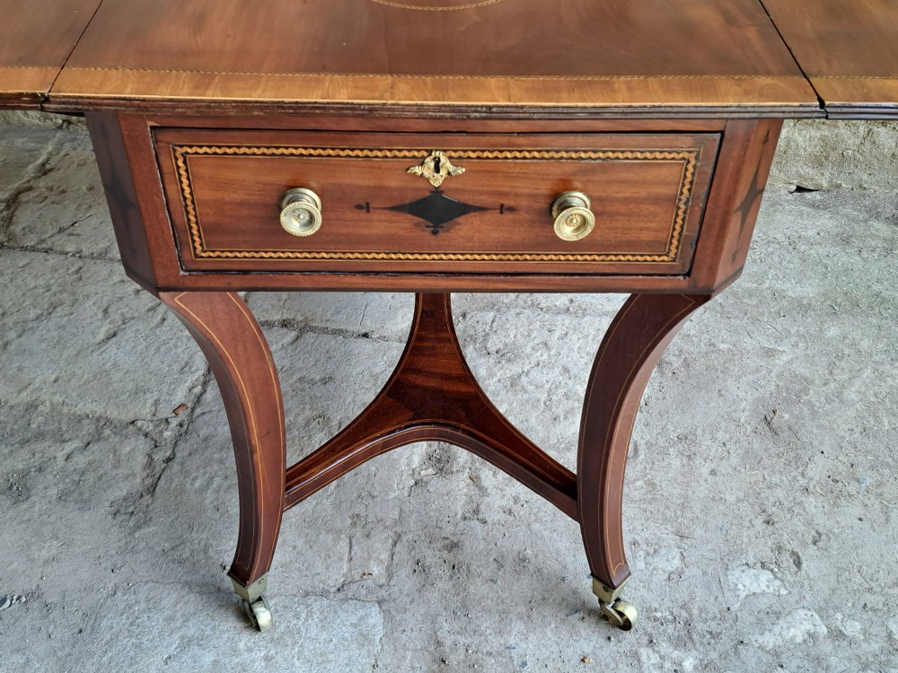 A Regency mahogany drop-leaf tea table featuring satinwood crossbanding, an oval marquetry motif, a single drawer with brass knobs, and splayed legs on castors. View IV
