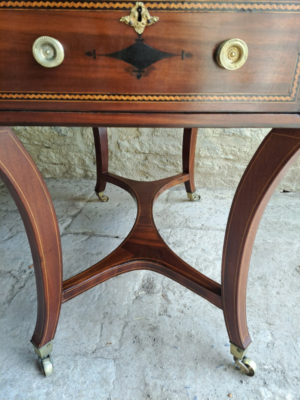 A Regency mahogany drop-leaf tea table featuring satinwood crossbanding, an oval marquetry motif, a single drawer with brass knobs, and splayed legs on castors. View V