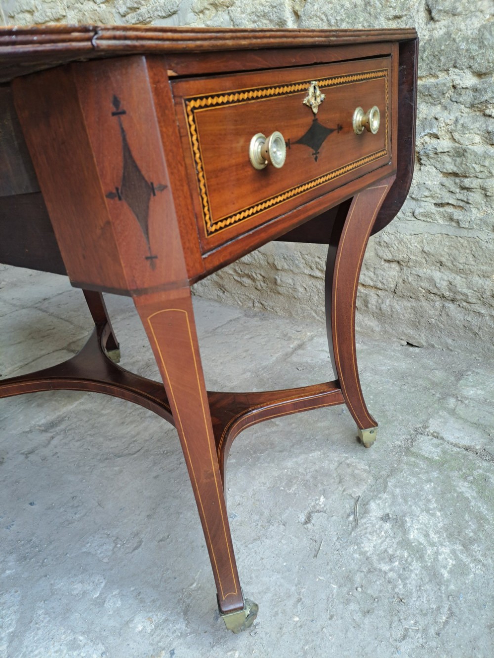 A Regency mahogany drop-leaf tea table featuring satinwood crossbanding, an oval marquetry motif, a single drawer with brass knobs, and splayed legs on castors. View VI