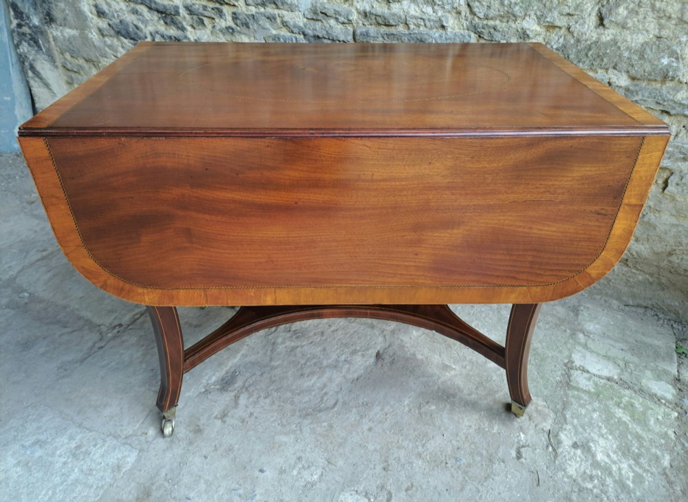A Regency mahogany drop-leaf tea table featuring satinwood crossbanding, an oval marquetry motif, a single drawer with brass knobs, and splayed legs on castors. View VIII