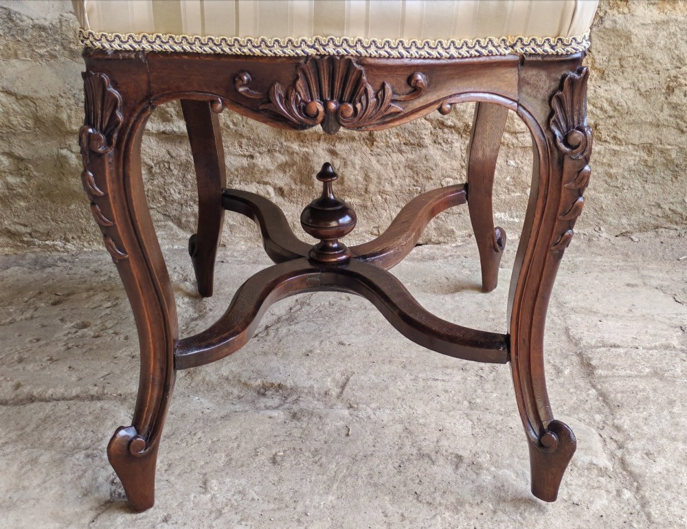 A carved dark mahogany Rococo style stool featuring a cream striped upholstered cushion, cabriole legs, carved shell motifs, and an X-stretcher with a turned finial. View III
