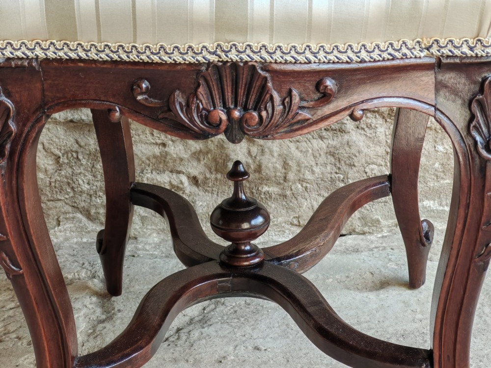 A carved dark mahogany Rococo style stool featuring a cream striped upholstered cushion, cabriole legs, carved shell motifs, and an X-stretcher with a turned finial. View IV