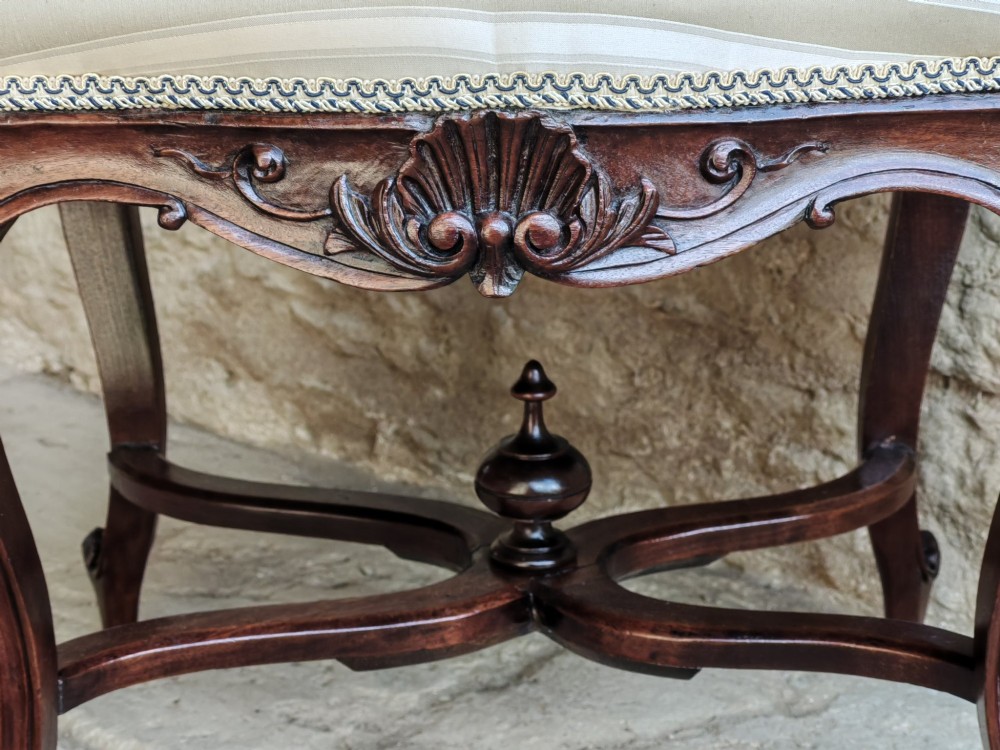 A carved dark mahogany Rococo style stool featuring a cream striped upholstered cushion, cabriole legs, carved shell motifs, and an X-stretcher with a turned finial. View V