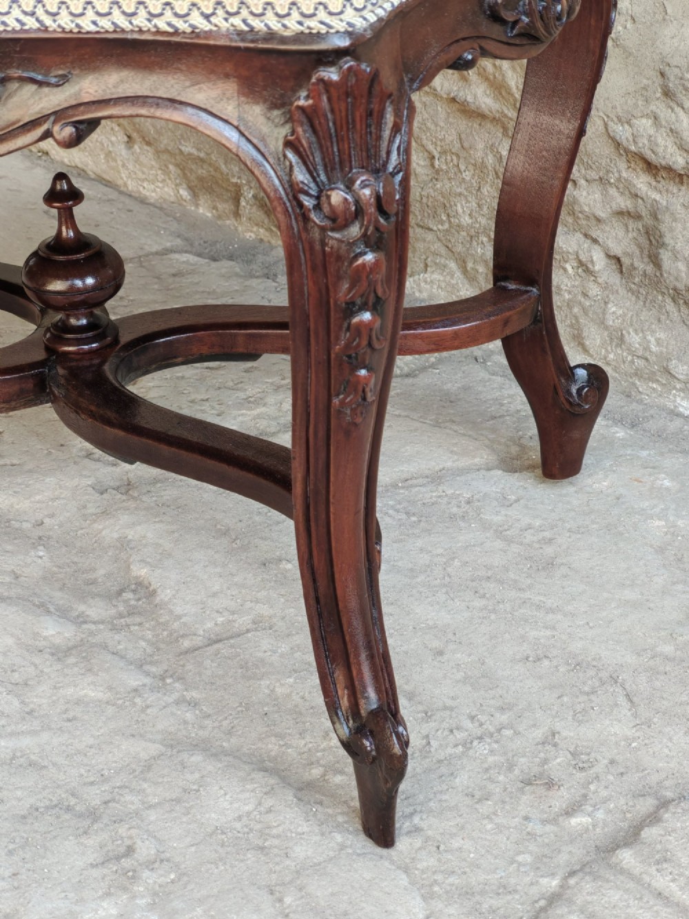 A carved dark mahogany Rococo style stool featuring a cream striped upholstered cushion, cabriole legs, carved shell motifs, and an X-stretcher with a turned finial. View VI