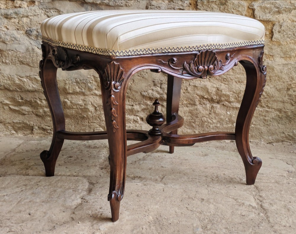 A carved dark mahogany Rococo style stool featuring a cream striped upholstered cushion, cabriole legs, carved shell motifs, and an X-stretcher with a turned finial.