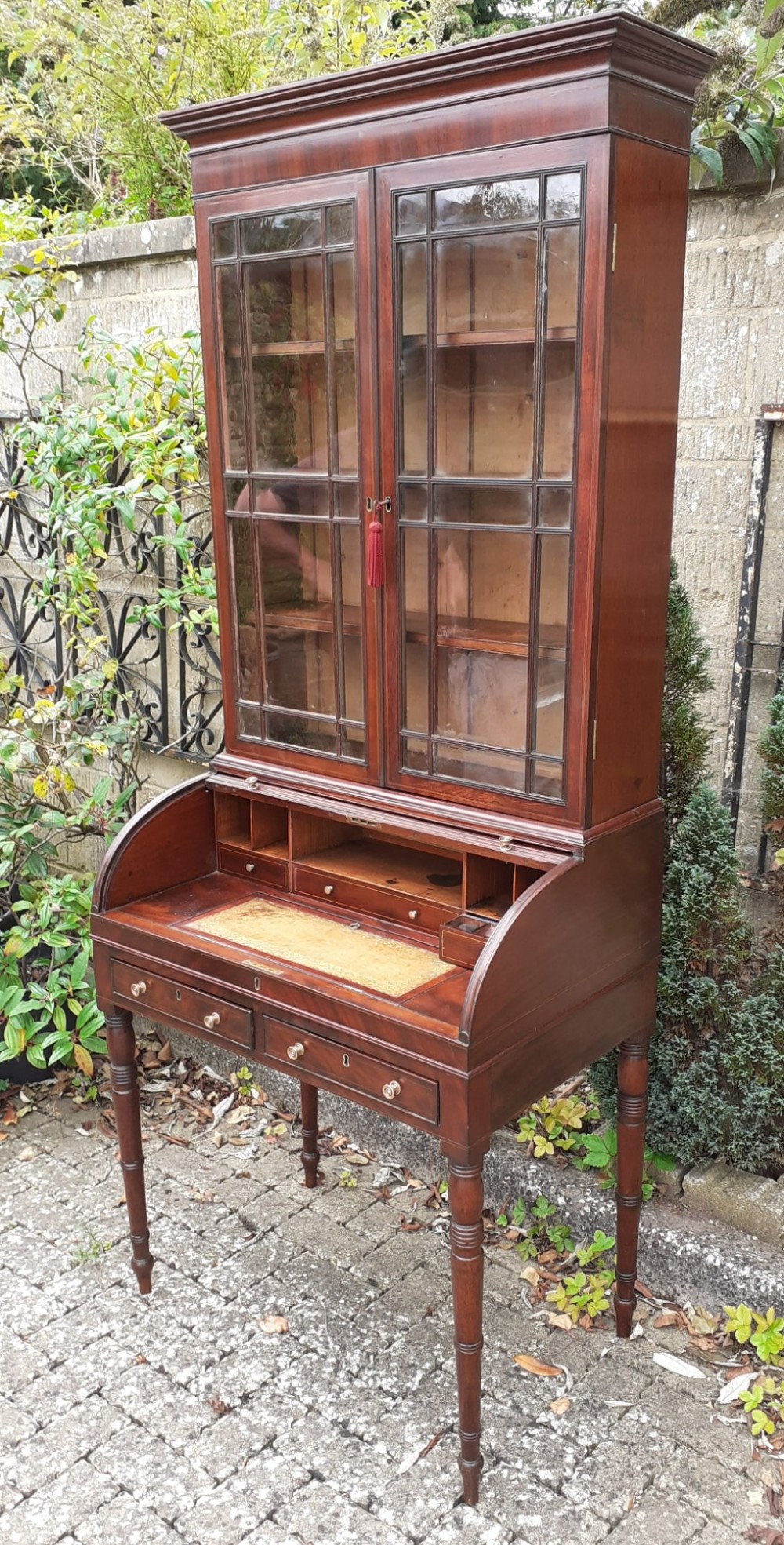 An antique mahogany bureau bookcase with a glazed upper door cabinet resting on a cylinder roll-top writing desk with turned legs.