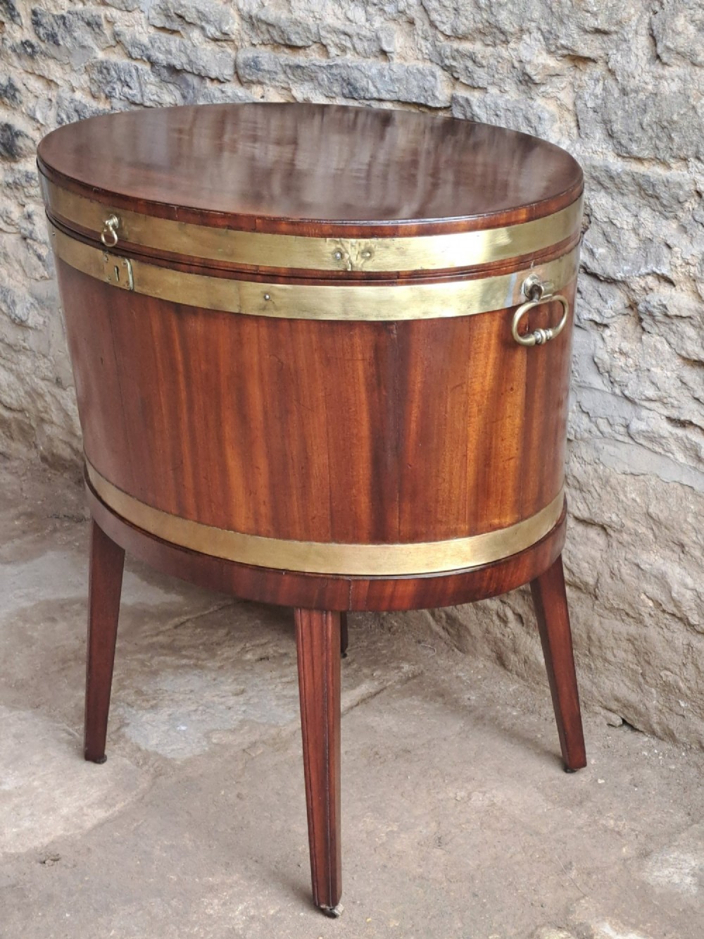 An 18th-century George III oval mahogany cellarette bound with brass straps and supported by four tapered legs.