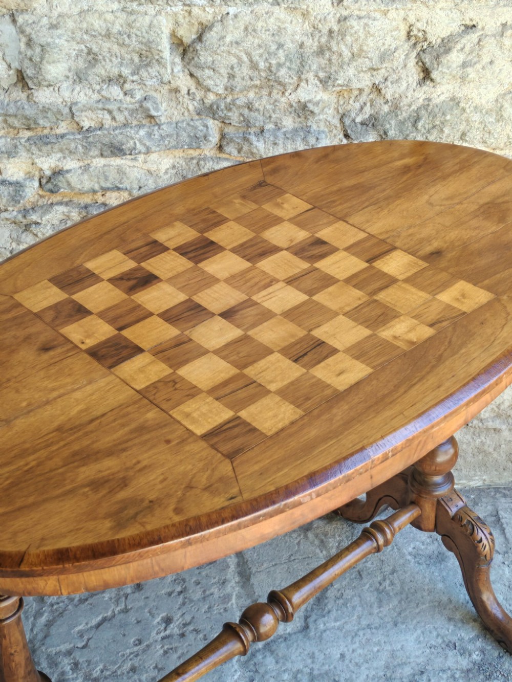 An oval walnut occasional table featuring an inlaid square chess board in the centre, supported by turned legs and a stretcher base. View V