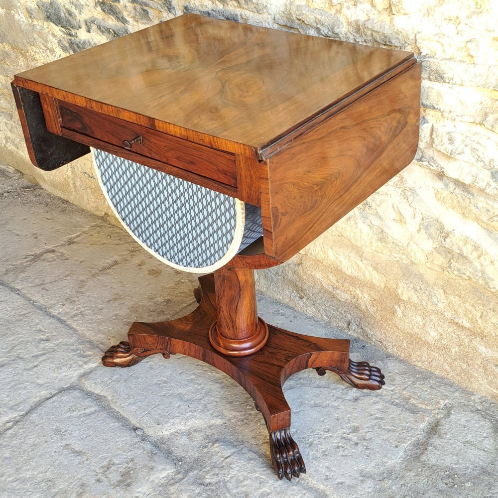 An antique rosewood drop-leaf sewing table with a central drawer, a suspended patterned fabric basket, a turned pedestal column, and carved claw feet.