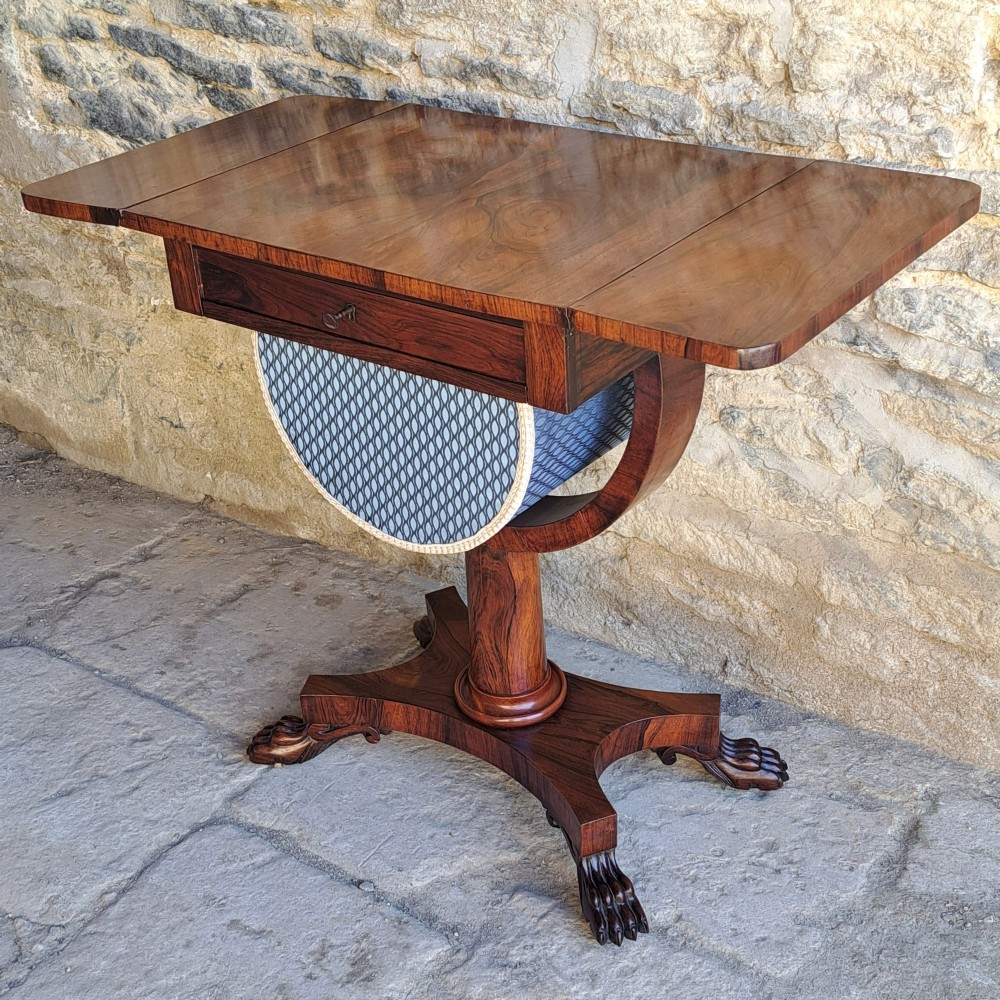 An antique rosewood drop-leaf sewing table with a central drawer, a suspended patterned fabric basket, a turned pedestal column, and carved claw feet. View II
