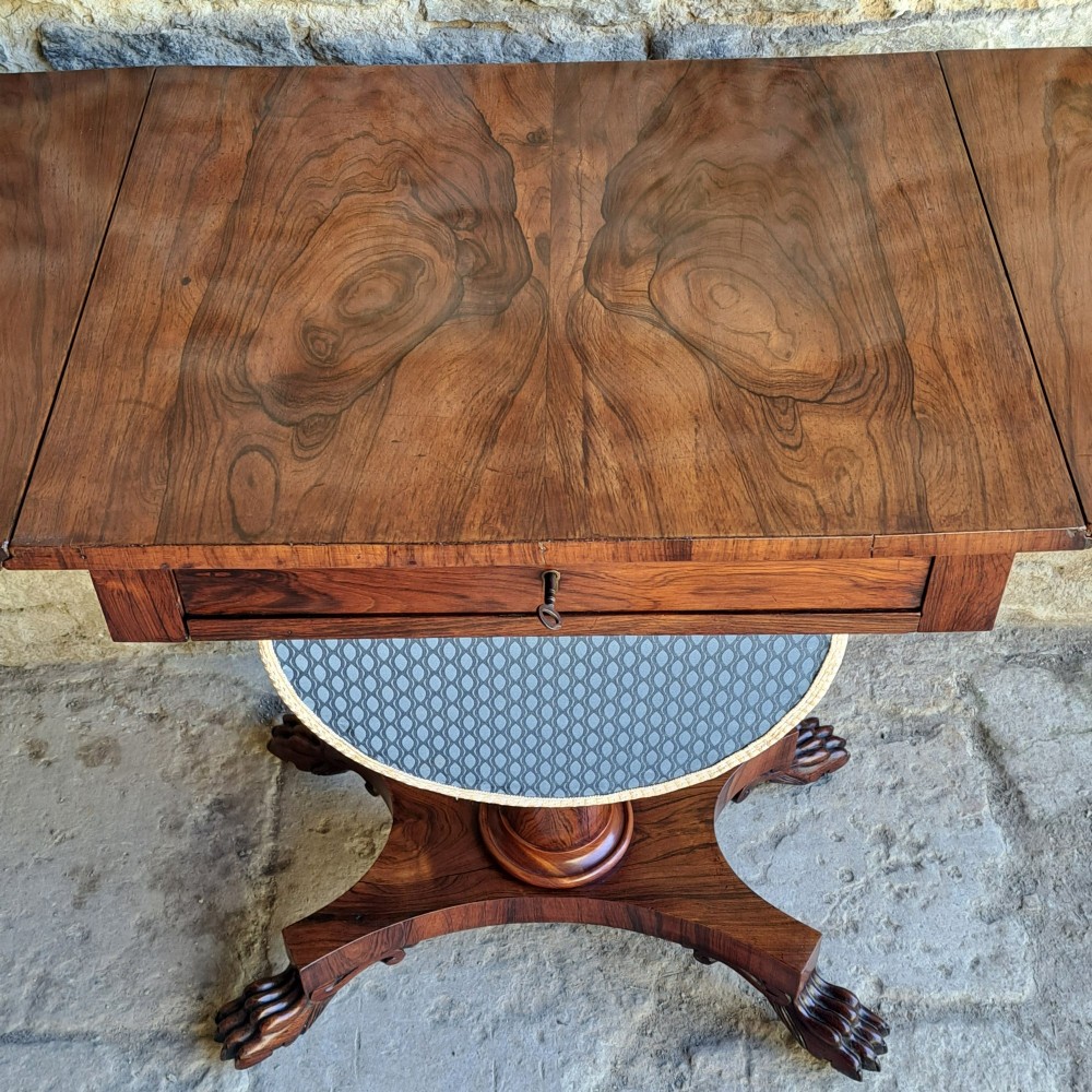 An antique rosewood drop-leaf sewing table with a central drawer, a suspended patterned fabric basket, a turned pedestal column, and carved claw feet. View IV