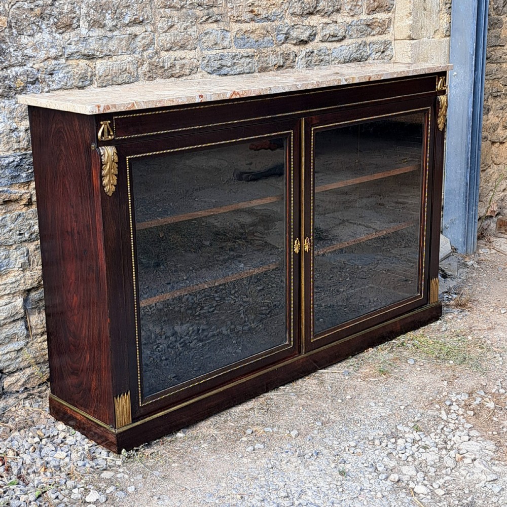 A low Regency rosewood glazed bookcase featuring a light marble top, twin glass doors framed in brass trim, and gilt-bronze ormolu corner accents.