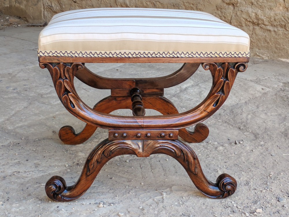 A Regency rosewood X-frame stool featuring intricately carved acanthus leaf scroll legs, a turned central stretcher, and a striped upholstered seat.