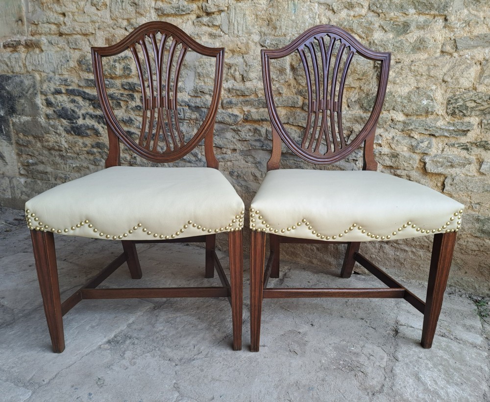 A pair of late 18th-century mahogany Hepplewhite dining chairs featuring carved shield backs and cream leather seats with brass stud detailing.