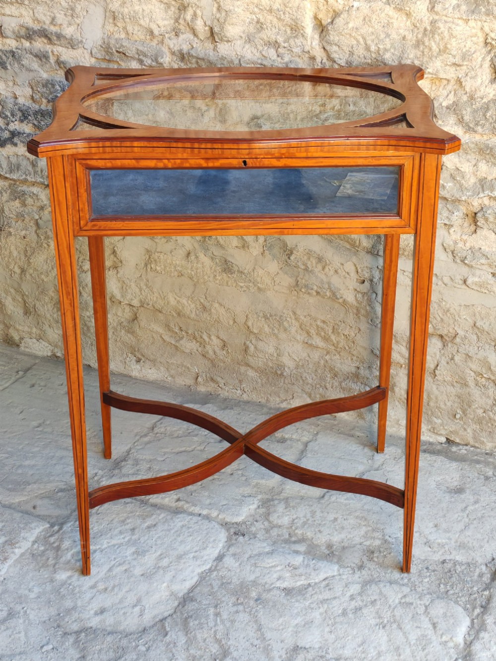 A late Victorian satinwood bijouterie table featuring an oval glazed top aperture, glass side panels, tapered square legs, and an X-form stretcher.