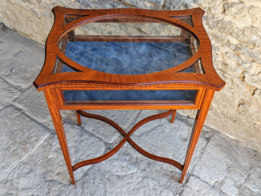 A late Victorian satinwood bijouterie table featuring an oval glazed top aperture, glass side panels, tapered square legs, and an X-form stretcher. View IV