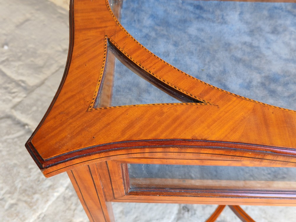 A late Victorian satinwood bijouterie table featuring an oval glazed top aperture, glass side panels, tapered square legs, and an X-form stretcher. View VI