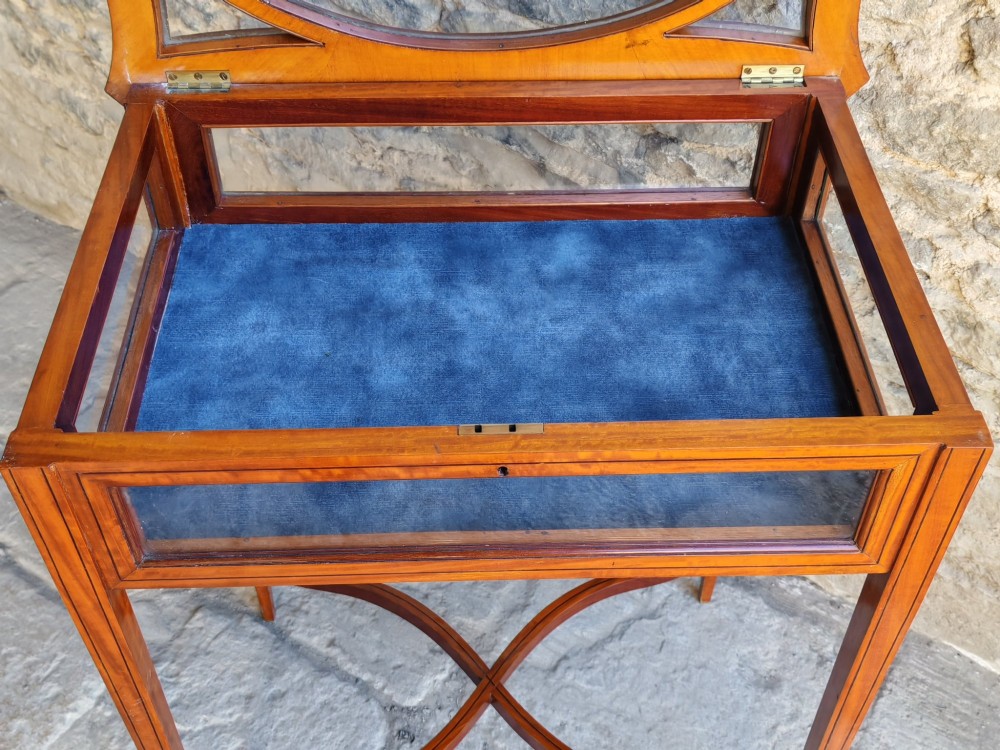 A late Victorian satinwood bijouterie table featuring an oval glazed top aperture, glass side panels, tapered square legs, and an X-form stretcher. View VII