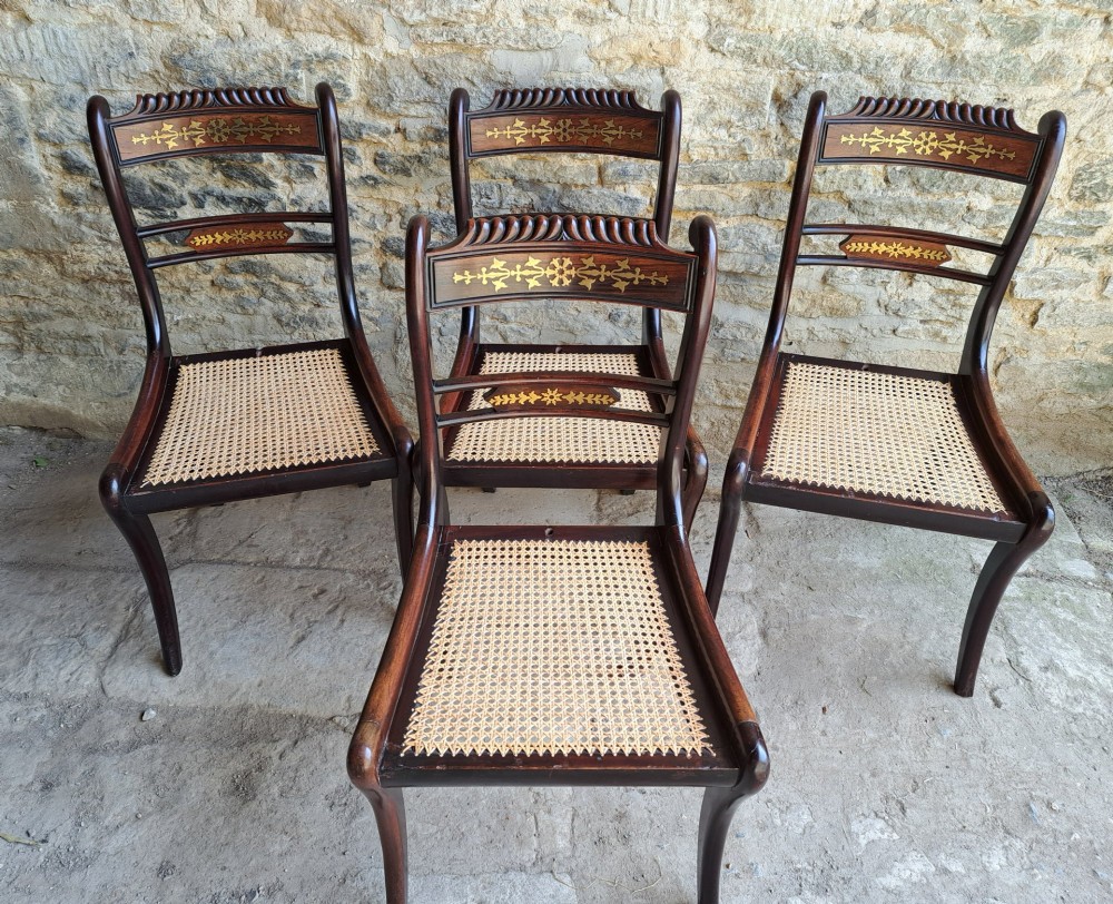 A set of four dark rosewood Regency dining chairs featuring intricate brass inlaid backrails, carved crests, hand-woven cane seats, and curved sabre legs.