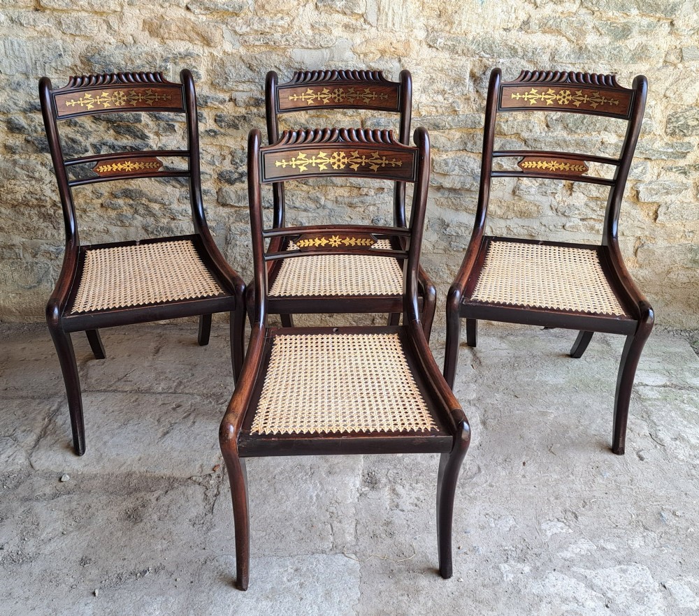 A set of four dark rosewood Regency dining chairs featuring intricate brass inlaid backrails, carved crests, hand-woven cane seats, and curved sabre legs. View II