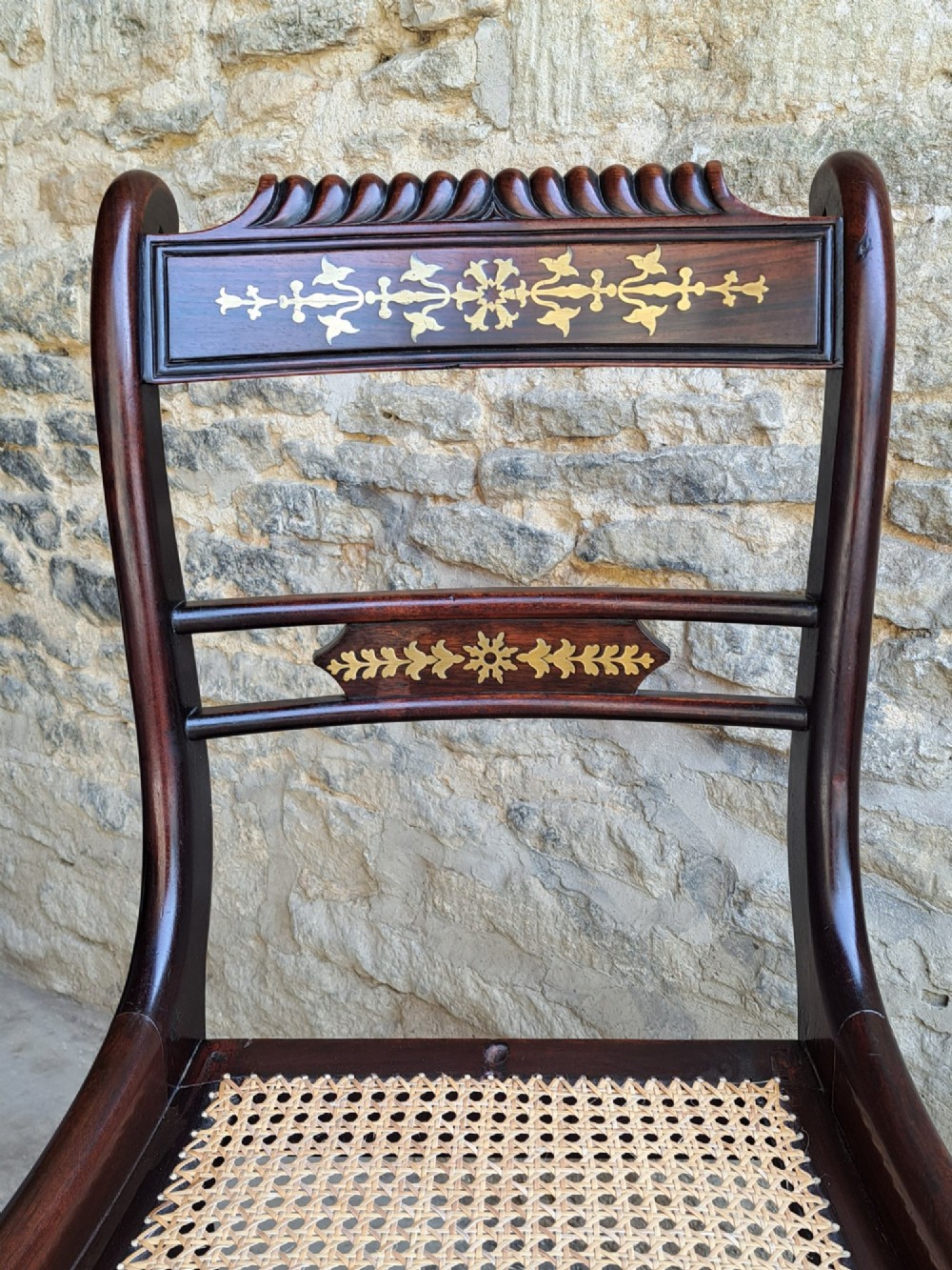 A set of four dark rosewood Regency dining chairs featuring intricate brass inlaid backrails, carved crests, hand-woven cane seats, and curved sabre legs. View V