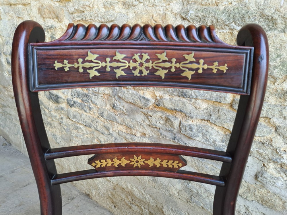 A set of four dark rosewood Regency dining chairs featuring intricate brass inlaid backrails, carved crests, hand-woven cane seats, and curved sabre legs. View VI